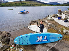 Lady in a wetsuit with a blue paddleboard standing at the foot of Briar Cottages slipway in to Loch Earn
