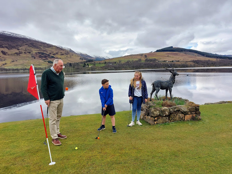 Boy in a blue jersey putts on Briar Cottages lawn with his sister and grandad watching. Loch Earn and Stan The Stag installation behindide putting a family