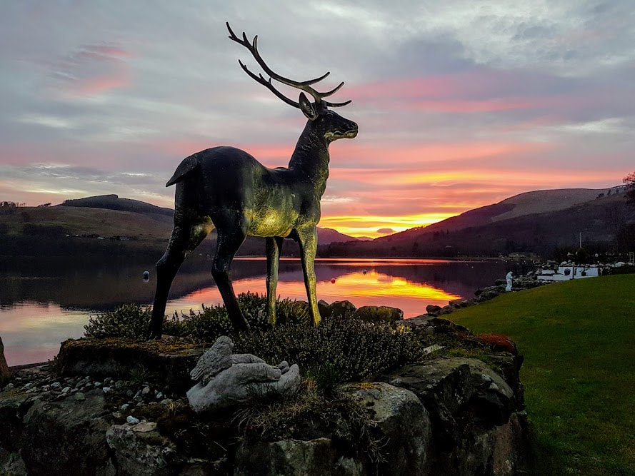 Stan the stag installation on a stony hillock with a pink and yellow sunset reflecting on Loch Earn behind