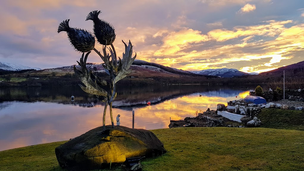 Blawn Wi The WInd double thistle headed metal sculpture against Loch Earn on a pink and yellow sunset evening 